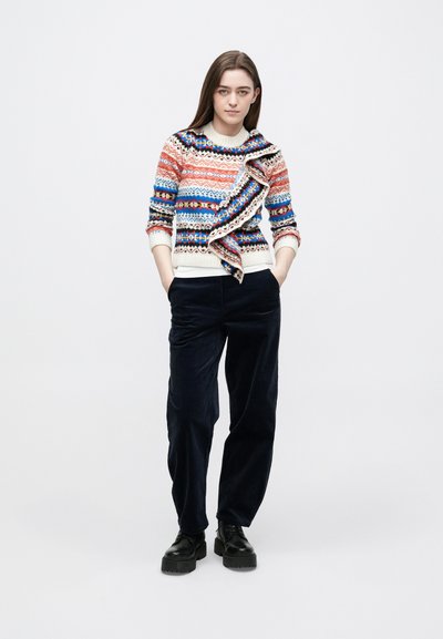 Adīts džemperis ar daudzkrāsainiem rakstiem sarkanā, zilā un krēmkrāsā, ar drapētu dizainu; kopā ar tumši ziliem korduroja biksēm un melniem apaviem.