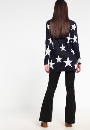 Femme aux longs cheveux raides portant un pull bleu marine avec des étoiles blanches, un pantalon noir évasé et des chaussures à talons marron, debout, vue de dos.
