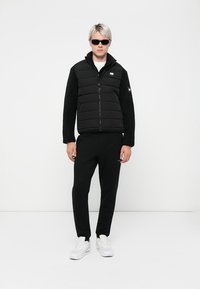 Sort puffer vest med en fleece-nederdel, lynlås foran og sidelommer, parret med sorte sweatpants og hvide sneakers.