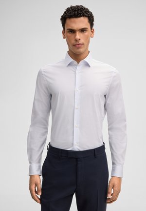 Mann mit lockigem Haar trägt ein hellgraues Hemd, das in eine dunkelblaue Hose gesteckt ist, und steht vor einem schlichten hellen Hintergrund.