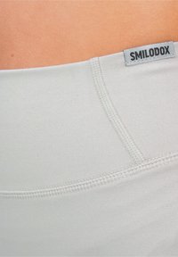 Leggings sportivi grigio chiaro con una texture liscia e elastica. Presentano una vita piatta e un'etichetta con la scritta "SMILODOX" sulla vita.