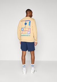 Beige sweatshirt with blue and orange geometric graphic on back, paired with navy shorts, white sneakers, and striped socks.