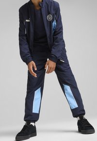 Survêtement bleu marine avec des accents bleu clair, comprenant une veste zippée et un pantalon à taille élastique. Inclut un logo sur la poitrine, design slim fit.