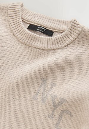 Light beige sweater with a ribbed crew neck, knit texture, and subtle 'NYC' print in dark grey. Brand label reads "NEXT, MADE IN CHINA."