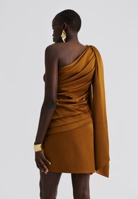 Vestido castanho assimétrico em tecido de cetim, com detalhes drapeados no corpete e uma capa fluida ao ombro. Acessórios dourados.