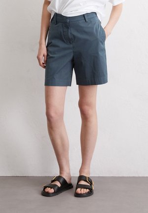 Persoon die donkergrijze knielange korte broek, wit ingestopt shirt en zwarte sandalen met gouden gespen draagt, staand tegen een effen muur.