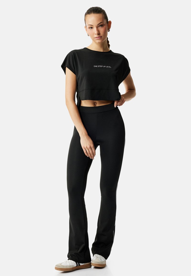 Donna in piedi con la mano sul fianco, indossa un crop top nero con la scritta "ONE STEP UP LEVEL", leggings neri e scarpe da ginnastica bianche su sfondo uniforme.