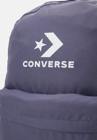 Mochila de color púrpura oscuro con un logo de estrella y chevron de Converse blanco impreso sobre un bolsillo frontal con cremallera.