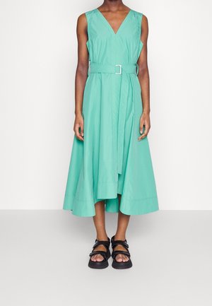 Robe midi sans manches vert menthe avec col en V et ceinture, portée avec des sandales noires à bouts ouverts sur une personne debout sur un fond uni.
