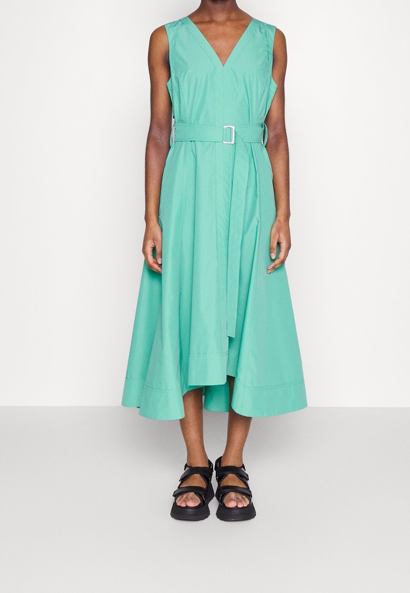 Robe midi sans manches vert menthe avec col en V et ceinture, portée avec des sandales noires à bouts ouverts sur une personne debout sur un fond uni.