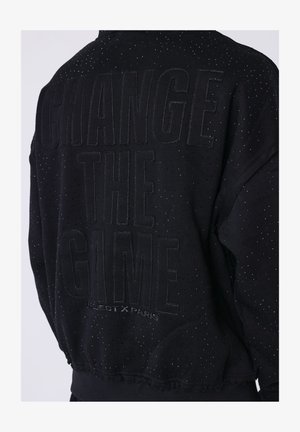 Sudadera negra con perforaciones sutiles y texto bordado "CAMBIA EL JUEGO" y en tamaño más pequeño "PROJECT X PARIS" en la parte trasera.