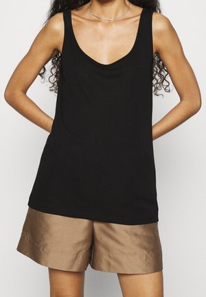 Kobieta w czarnej, bezrękawnikowej bluzce z zaokrąglonym dekoltem i jasnobrązowych krótkich spodenkach do kolan, z rękami spoczywającymi za plecami.