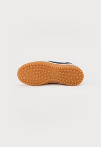 Semelle de chaussure avec une bande de roulement en caoutchouc orange présentant des crampons carrés et un logo Nike au centre sur un fond blanc uni.