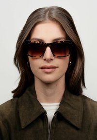 Occhiali da sole tartarugati con montatura oversized e angolare. Lenti scure con gradiente. Il modello ha lunghi capelli castani e indossa una giacca in suede verde oliva.