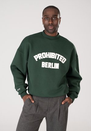 Mies, joka pukeutunut tummanvihreään collegepaitaan, jossa lukee valkoisella "PROHIBITED BERLIN", seisoo kädet taskuissa yksinkertaisen vaalean taustan edessä.