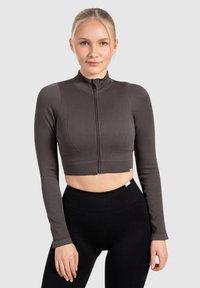Bruine geribbelde cropped zip-up top met lange mouwen en hoge kraag; gecombineerd met zwarte high-waisted leggings met een gladde textuur.