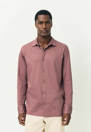 Mann steht vor einem weißen Hintergrund und trägt ein langärmliges, rosafarbenes Hemd und hellbeige Hosen.