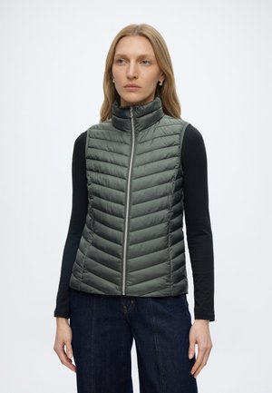 Frau trägt graue gesteppte ärmellose Jacke über schwarzem Langarmshirt und dunkelblauen Jeans, stehend vor einfachem weißem Hintergrund.