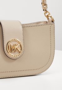 Sac en bandoulière en synthétique beige avec une forme arrondie, une surface texturée, un matériel logo doré et une chaîne en bandoulière. Les détails de couture sont visibles.