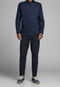 Camicia a maniche lunghe blu navy con bottoni, pantaloni a quadri testurizzati grigio antracite, sneakers grigie con accenti blu e giallo.