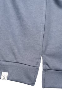 Szara bawełniana tkanina o gładkiej fakturze, z widocznymi szwami. Dolna część ma rozcięcie i małą metkę z logo.