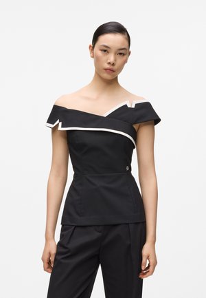 Mujer con un top negro de hombros descubiertos con ribete blanco y pantalones negros, de pie contra un fondo claro y sencillo.