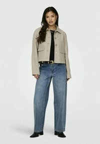 Casaco cropped bege claro com botões grandes e dois bolsos frontais, combinado com jeans de ganga azul largos e botas pretas até ao tornozelo.