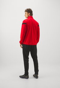 Veste de sport rouge avec des accents noirs, assortie à un pantalon noir et des chaussures de sport noires. La veste a un col montant et des manches longues.