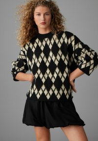 Pull en argyle noir et crème avec un col rond, des manches oversize et une finition texturée, associé à une jupe noire à volants.