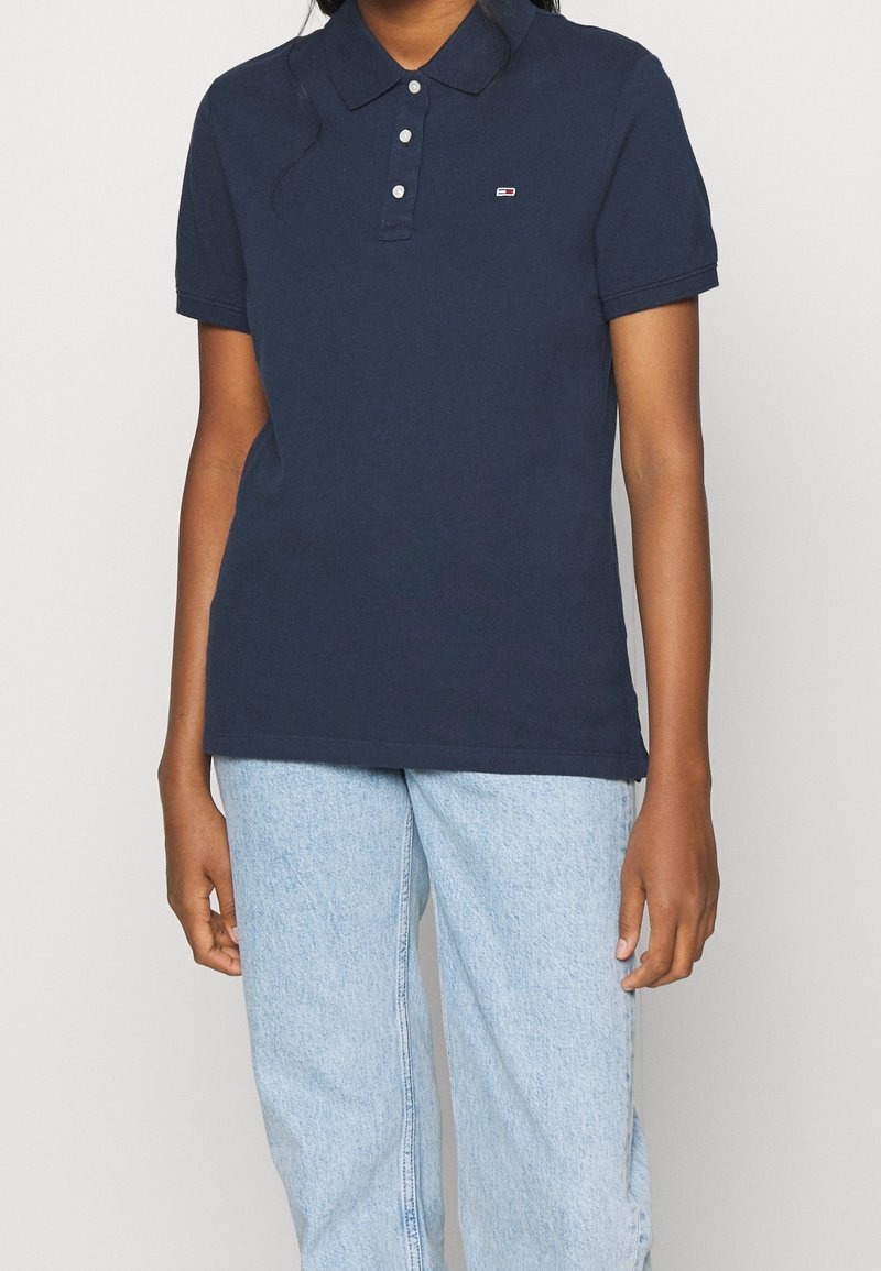 Mörkblå piké med krage, tre knappar och en liten logotyp. Återfinns tillsammans med ljusblå denimjeans med rak benkomfort.