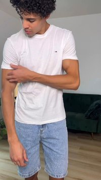 Jeune homme aux cheveux bouclés portant un t-shirt blanc et un short en jean, ajustant sa manche gauche à l'intérieur près d'un canapé vert foncé.