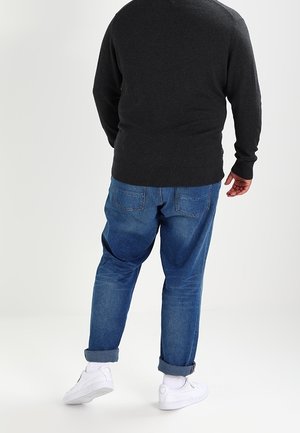 Mann trägt einen dunklen Pullover, hochgekrempelte blaue Jeans, weiße Socken und weiße Turnschuhe, steht mit dem Rücken zum Betrachter vor einem einfachen hellen Hintergrund.