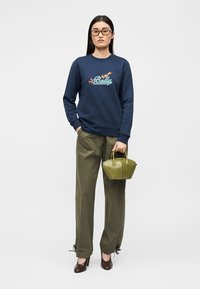 Sweat-shirt bleu marine avec broderie "Bally" multicolore, pantalon vert olive et petit sac à main olive ; le mannequin porte des chaussures à talons marron et des lunettes.