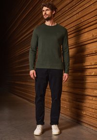 Camiseta de manga larga en verde oscuro hecha de tela suave, combinada con pantalones azul marino y zapatillas blancas. Presenta un clásico cuello redondo.