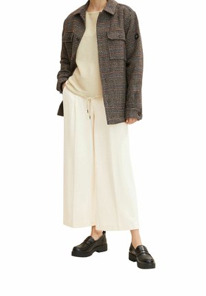 Person trägt eine braun karierte Jacke, einen beigen Pullover, lockere cremefarbene Hosen und schwarze Loafer und steht mit den Händen in den Taschen.