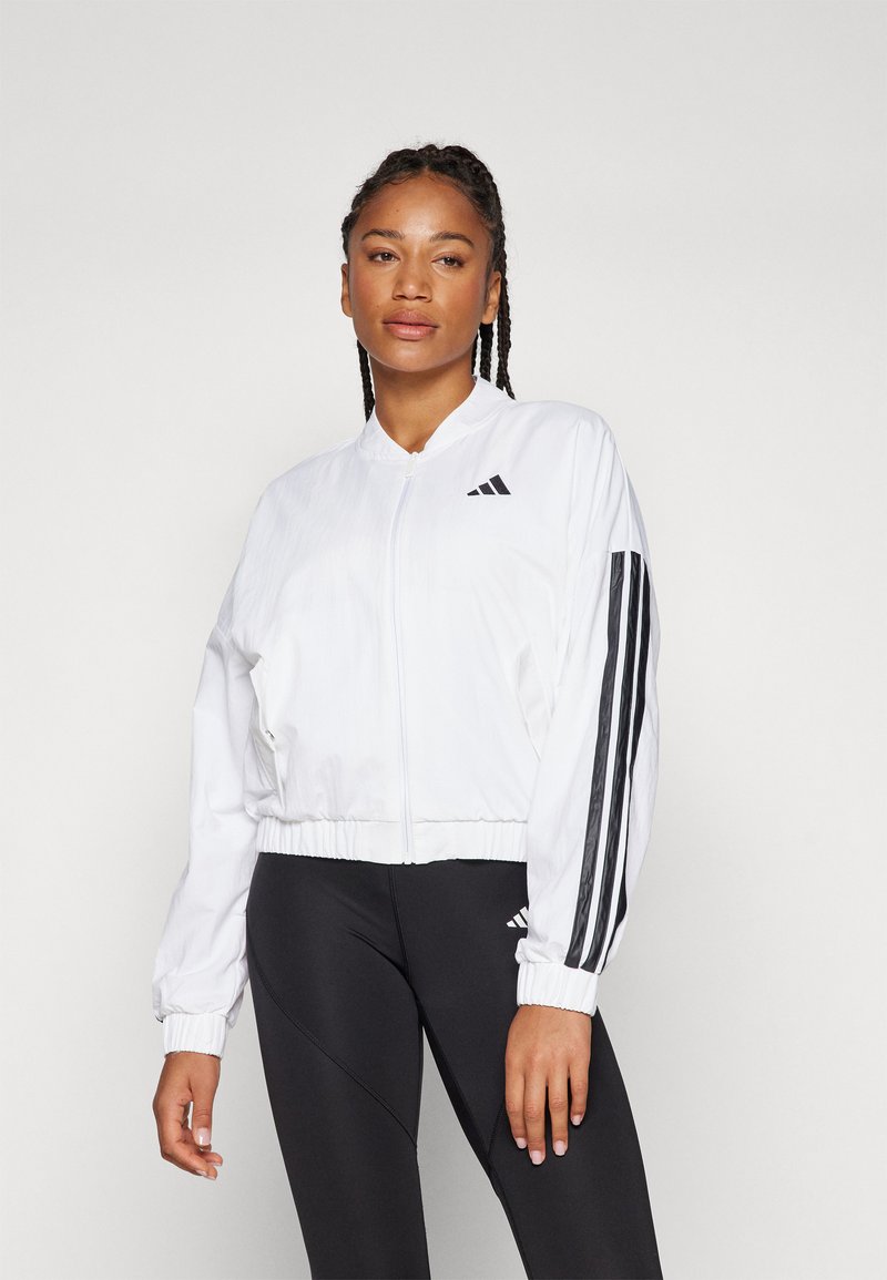 Bílá zipová bunda s ribbeným límcem a manžetami, se třemi černými pruhy podél rukávů a logem Adidas na levém prsu.