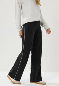 Pantalones de pierna ancha negros con piping blanco a los lados, hechos de una tela suave. Combinados con una sudadera gris y zapatillas deportivas de color claro.