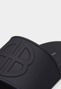 Μαύρη σαγιονάρα με ανάγλυφο μεγάλο διαπλεκόμενο λογότυπο "B" στο λουράκι, λεία υφή και ορατή ραφή.