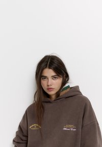 Hnědá oversized mikina s klokaní kapsou. Obsahuje žluté a bílé vyšívané nápisy. Kapuce je vyložená zeleno-bežovým kostkovaným vzorem.