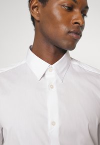 Weißes Hemd mit Knopfleiste, klassischem Kragen, glatter Textur und subtiler Satin-Optik, mit drei sichtbaren Knöpfen nahe dem Ausschnitt.