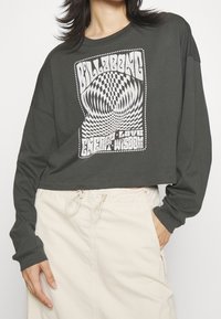 Persona che indossa un crop top lungo scuro con la scritta "Billabong" e un design a scacchi distorto, abbinato a una gonna di colore chiaro.