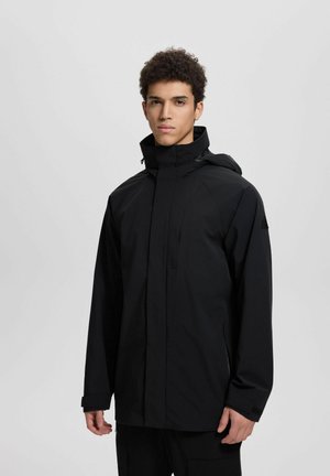 Młody mężczyzna z krótkimi kręconymi włosami, ubrany w czarny płaszcz z kapturem, z zamkiem błyskawicznym i rzepami na mankietach, stojący na jednolitym jasnoszarym tle.