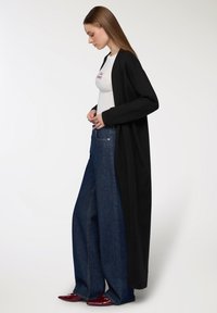 Cardigan lungo nero sopra una maglietta bianca, abbinato a jeans blu a gamba larga e scarpe rosse, che mette in risalto una silhouette rilassata e stratificata.