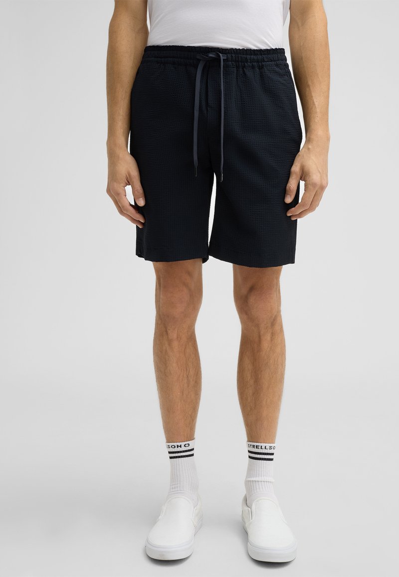 Marineblauwe shorts met een elastische tailleband en trekkoord. Geweven stof, knielange snit, gecombineerd met witte instappers en sokken.