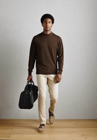 Bruine gebreide trui, crèmekleurige broek en bruine schoenen. Draagt een zwarte tas met zilveren accenten. Eenvoudig, gestroomlijnd ontwerp. Houten vloer.