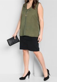 Top sin mangas en verde oliva con escote en V, acompañado de una falda negra hasta la rodilla y zapatos de tacón negro. Clutch negro en la mano.