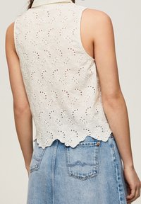 Top sans manches blanc avec un motif floral brodé, un ourlet festonné et une fermeture à bouton au col, associé à une jupe en denim.