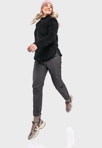 Schwarze Fleecejacke mit Reißverschluss, hohem Kragen und seitlichen Schlitzen. Graue Stretchhose und beiges Paar Sneaker mit rosa Schnürsenkeln. Rosa Mütze.