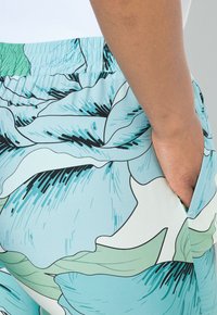 Pantalons à taille élastique avec un motif floral, présentant des feuilles bleu clair et vert sur un fond blanc. Poche latérale visible.