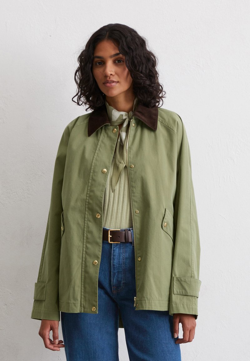 Olive green lightweight jacket with a brown collar, zip and snap closures, and side pockets, worn over a light blouse and blue jeans.